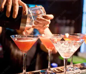A bartender pouring a red cocktail from a shaker into a martini glass, with other garnished cocktails lined up on the bar.