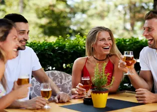 A group of four friends laughing and drinking beer at an outdoor table outdoors.