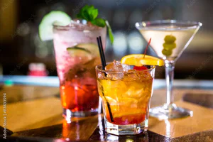 A tall red cocktail, an orange cocktail, and a martini with green olives and an orange slice on a wooden bar counter at The Yard in Seneca, SC.