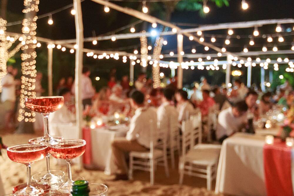 A nighttime outdoor wedding reception at The Yard in Seneca, SC with a string-lit canopy, guests seated at tables, and a champagne cocktail tower in the foreground.