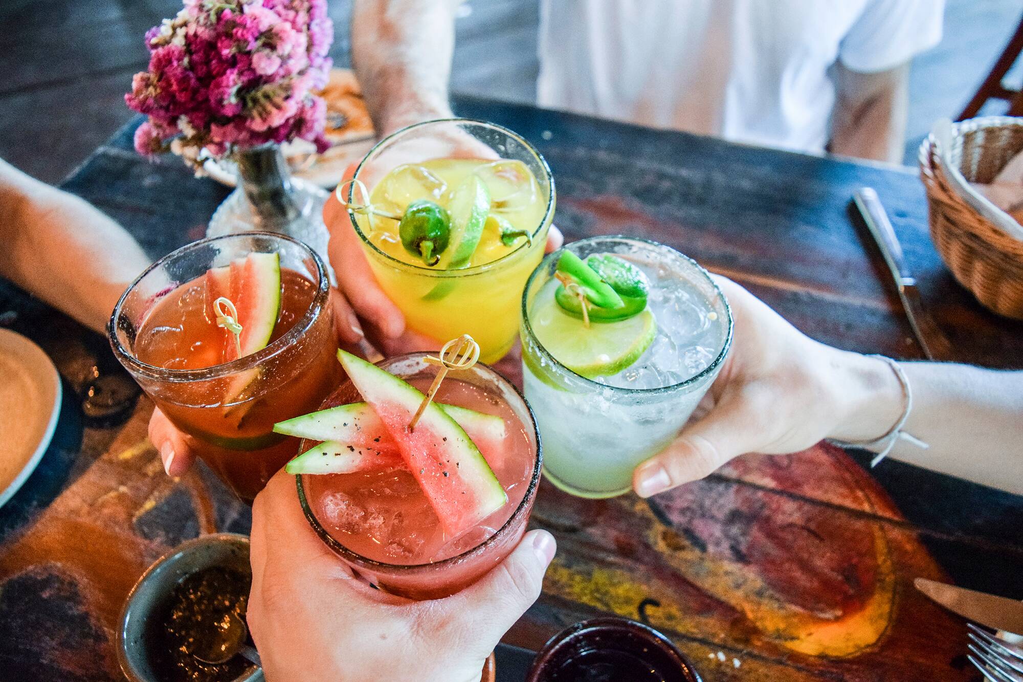 Four people toasting with assorted cocktails garnished with watermelon slices, lime wedges, and jalapeños over a wooden table.