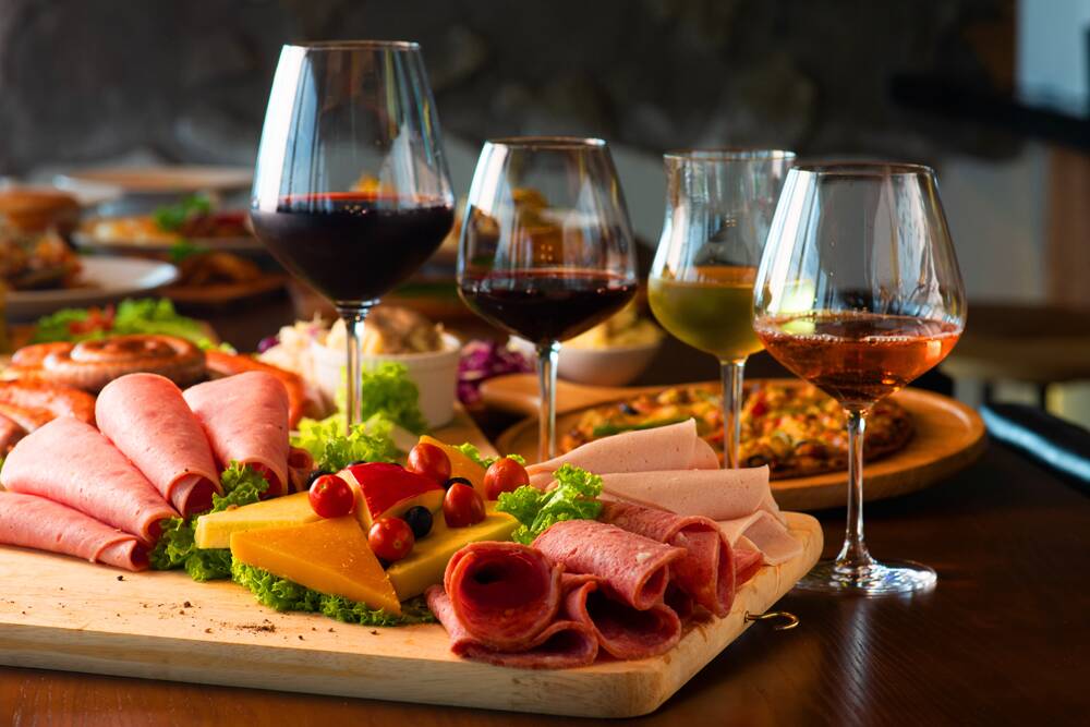 Platter with assorted meats, cheeses, cherry tomatoes, and four glasses of wine on a wooden board.