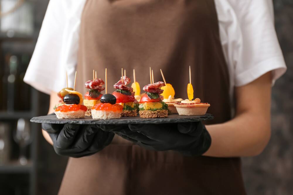 A person in an apron and black gloves holding a slate serving tray with assorted canapés and small tartlets at The Yard in Seneca, SC.