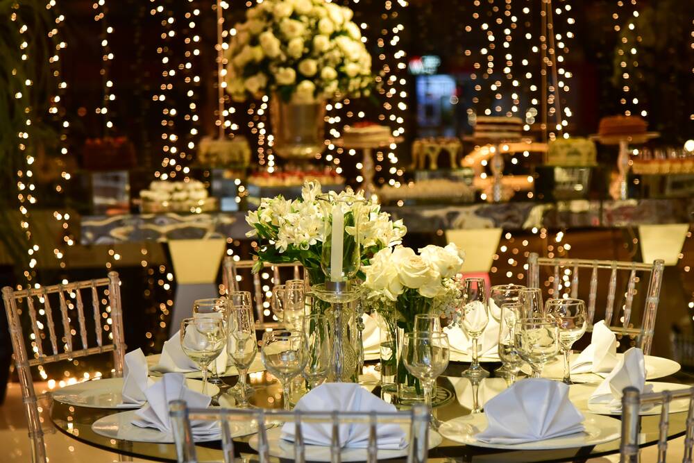 Elegant event table setup with white floral centerpieces, glassware, and candles with string lights in the background.