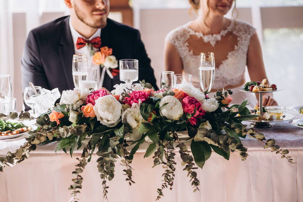 A bride and groom seated at a wedding reception table decorated with a large floral centerpiece and champagne flutes.