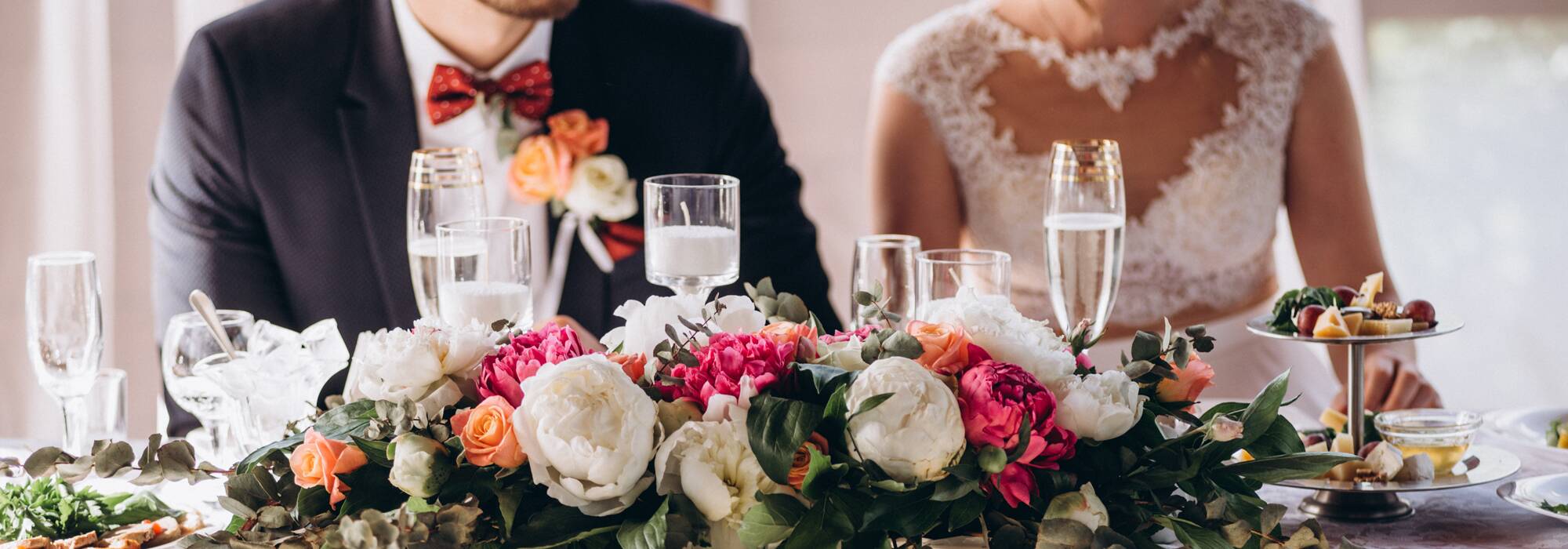 A bride and groom seated at a wedding reception table decorated with a large floral centerpiece and champagne flutes.